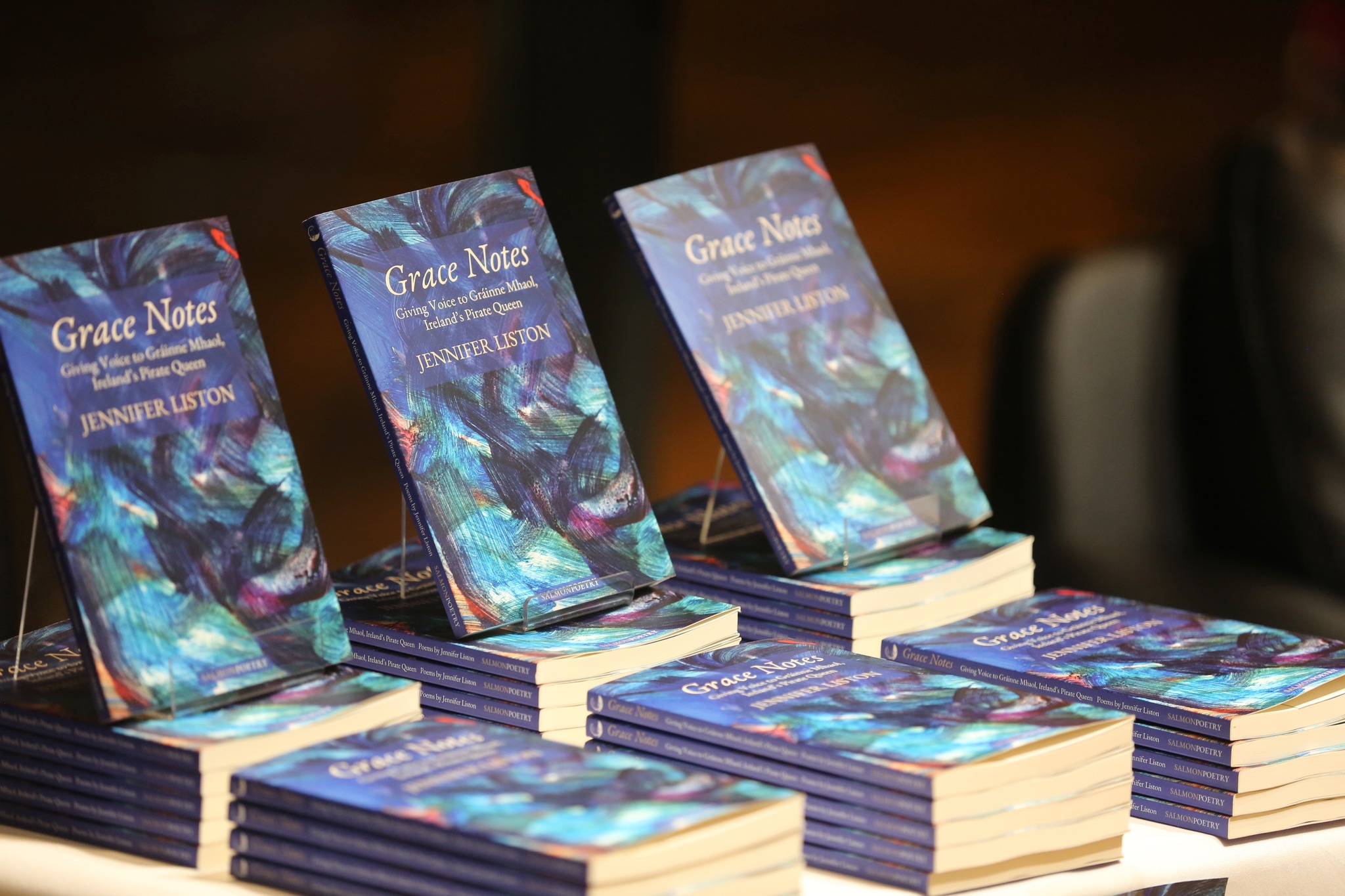 A display table with multiple stacks of Jennifer Liston's book, <em>Grace Notes: Giving Voice to Gráinne Mhaol, Ireland’s Pirate Queen</em>. Several copies are propped up to showcase the cover, which features a vibrant abstract design in blue, green, and purple tones. The books are neatly arranged, creating an inviting setup for a book signing or sale event. This ALT-text was generated by ChatGPT.