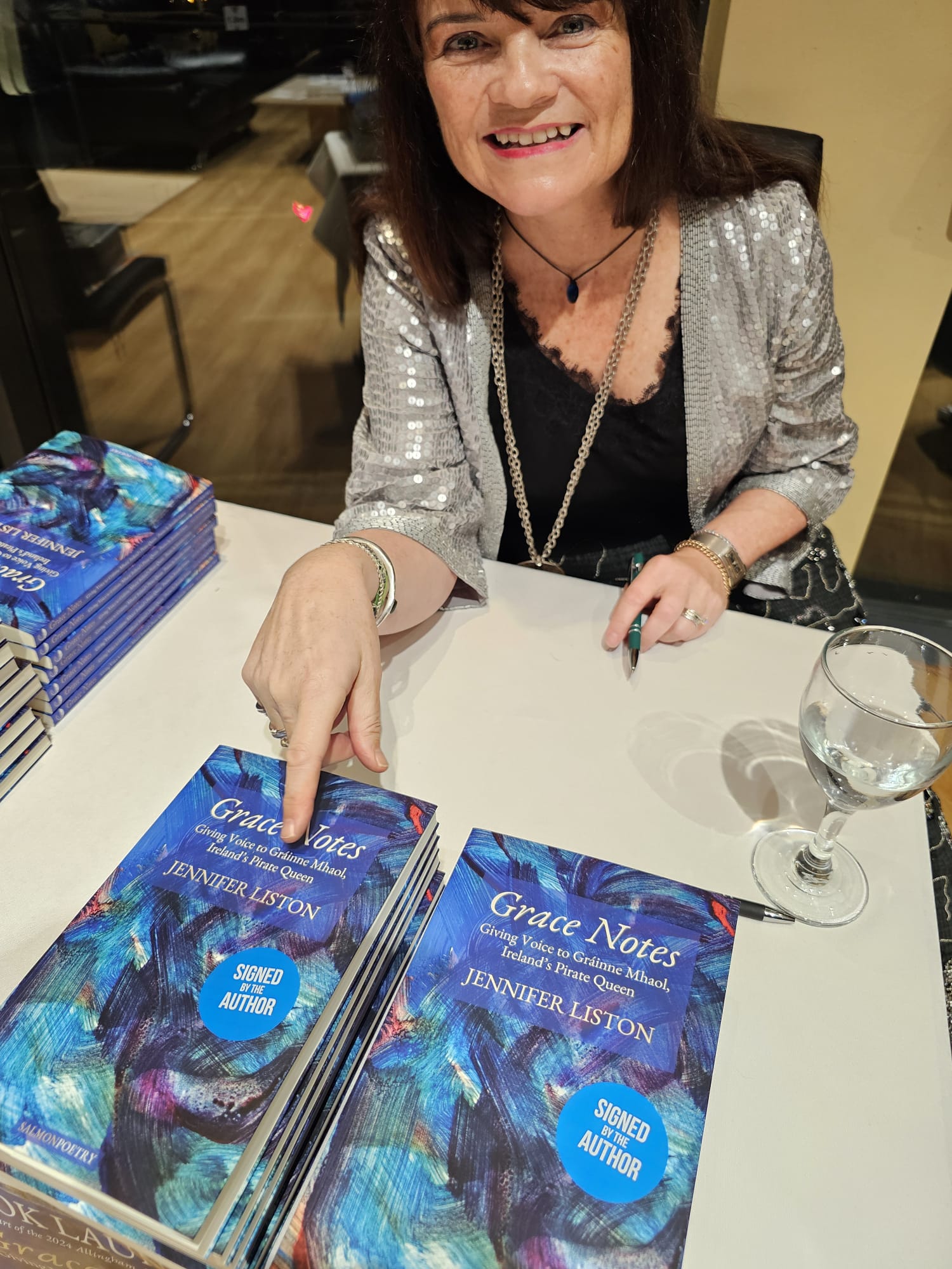 Author Jennifer Liston at a book signing event, smiling at the camera while pointing to a stack of her book, <em>Grace Notes: Giving Voice to Gráinne Mhaol, Ireland’s Pirate Queen</em>. She is wearing a sparkly silver jacket over a black top and a necklace. The books on the table have a blue and green abstract cover with a sticker saying 'Signed by the Author.' A wine glass sits nearby on the table. This ALT-text was generated by ChatGPT.