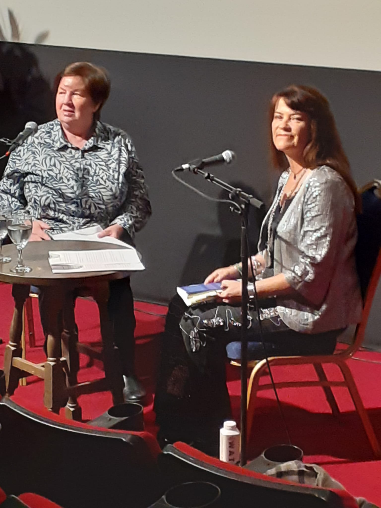 Author Jennifer Liston on stage seated next to Win McNulty at a small table with microphones in front of them. Jennifer, wearing a sparkly silver jacket, holds a copy of <em>Grace Notes: Giving Voice to Gráinne Mhaol, Ireland’s Pirate Queen</em> in her lap and looks toward the audience. The setting is a theatre with red carpeting and seating in the foreground. This ALT-text was generated by ChatGPT, then edited.